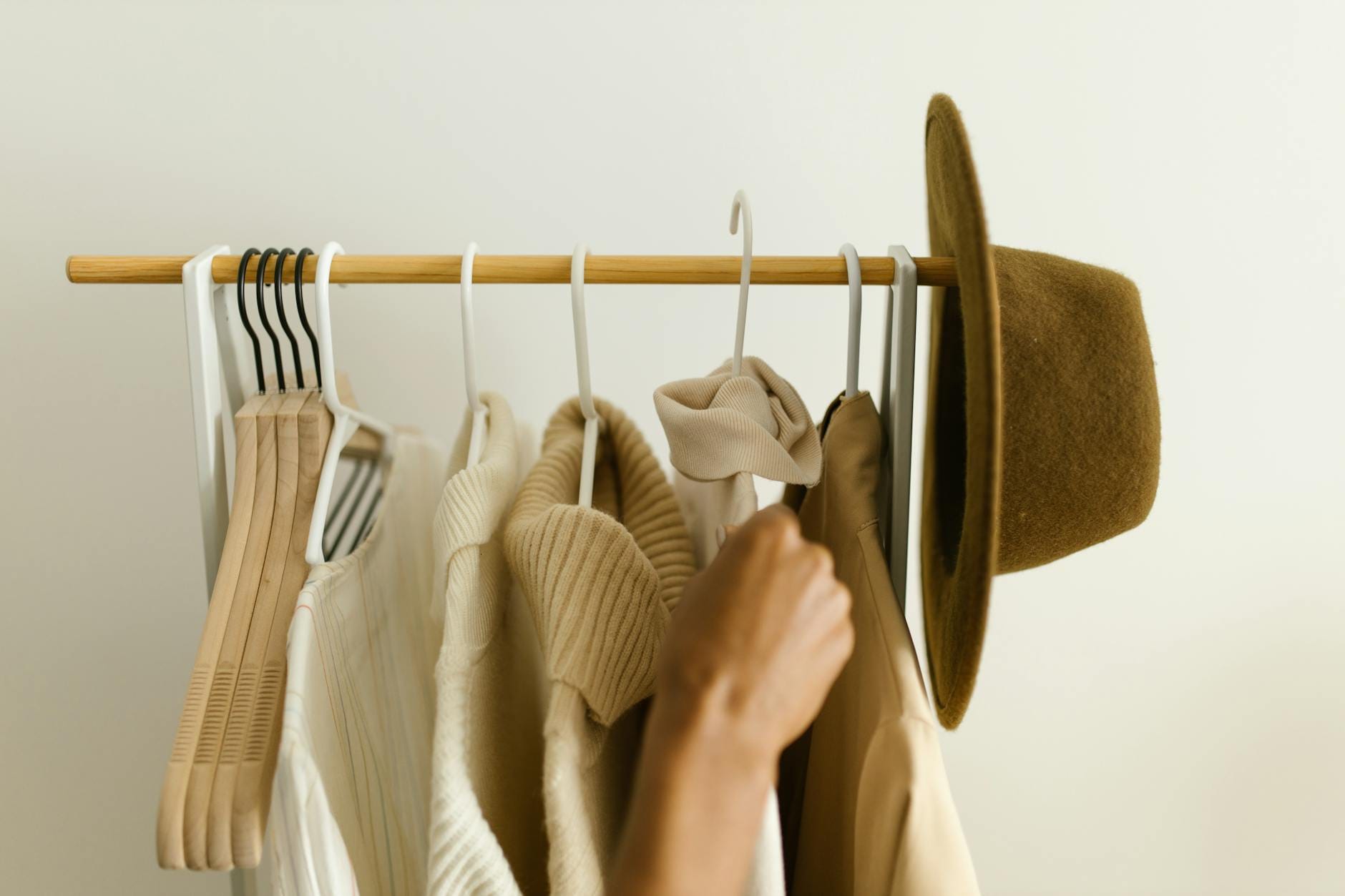 Jak kupować mniej, a lepiej: praktyczny przewodnik po slow fashion dla zabieganych Ręka sięga po beżowe i brązowe ubrania wiszące na wieszaku